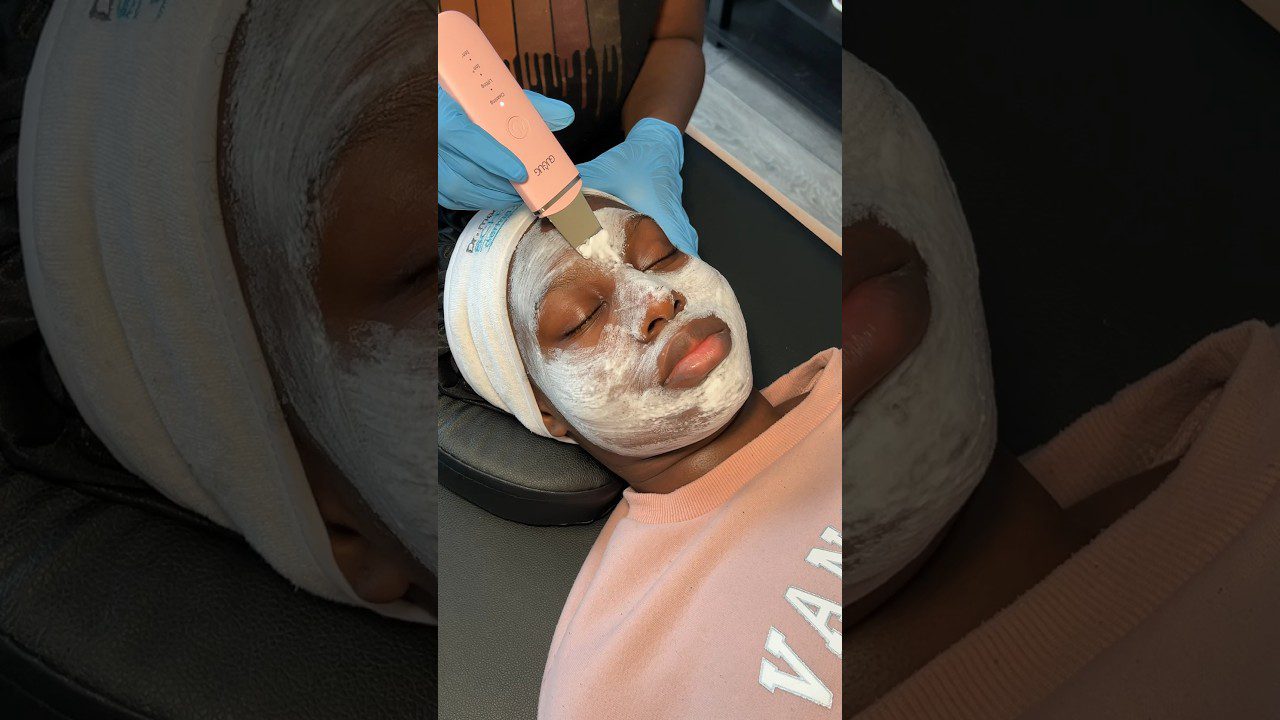 Moteris su veido kauke gulinti, kosmetologė su pirštinėmis šveičia odą ultragarsiniu šepetėliu