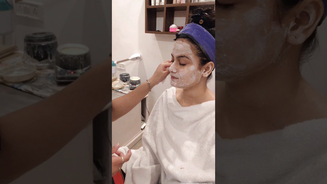 Moteris su veido kauke ir rankšluosčiu, kosmetologė tepasi kremą ant jos veido.