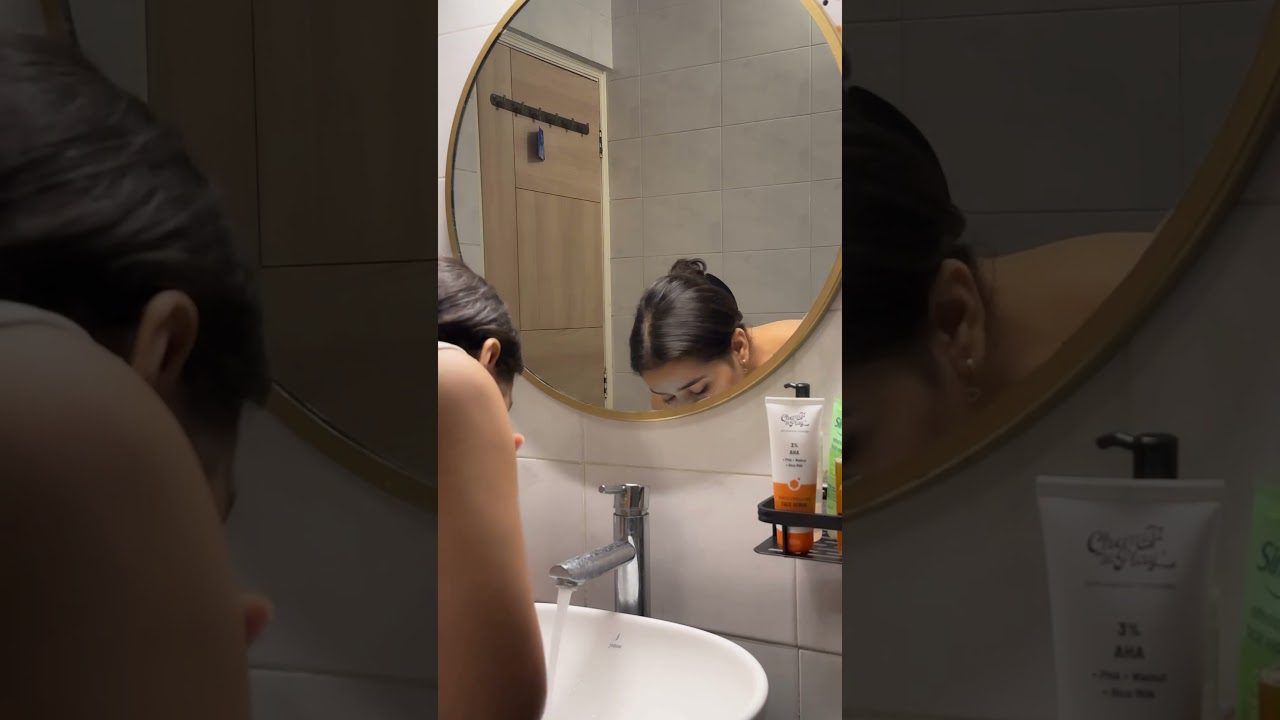 Moteris lenkiasi prie kriauklės ir plauna veidą prieš apvalų veidrodį.