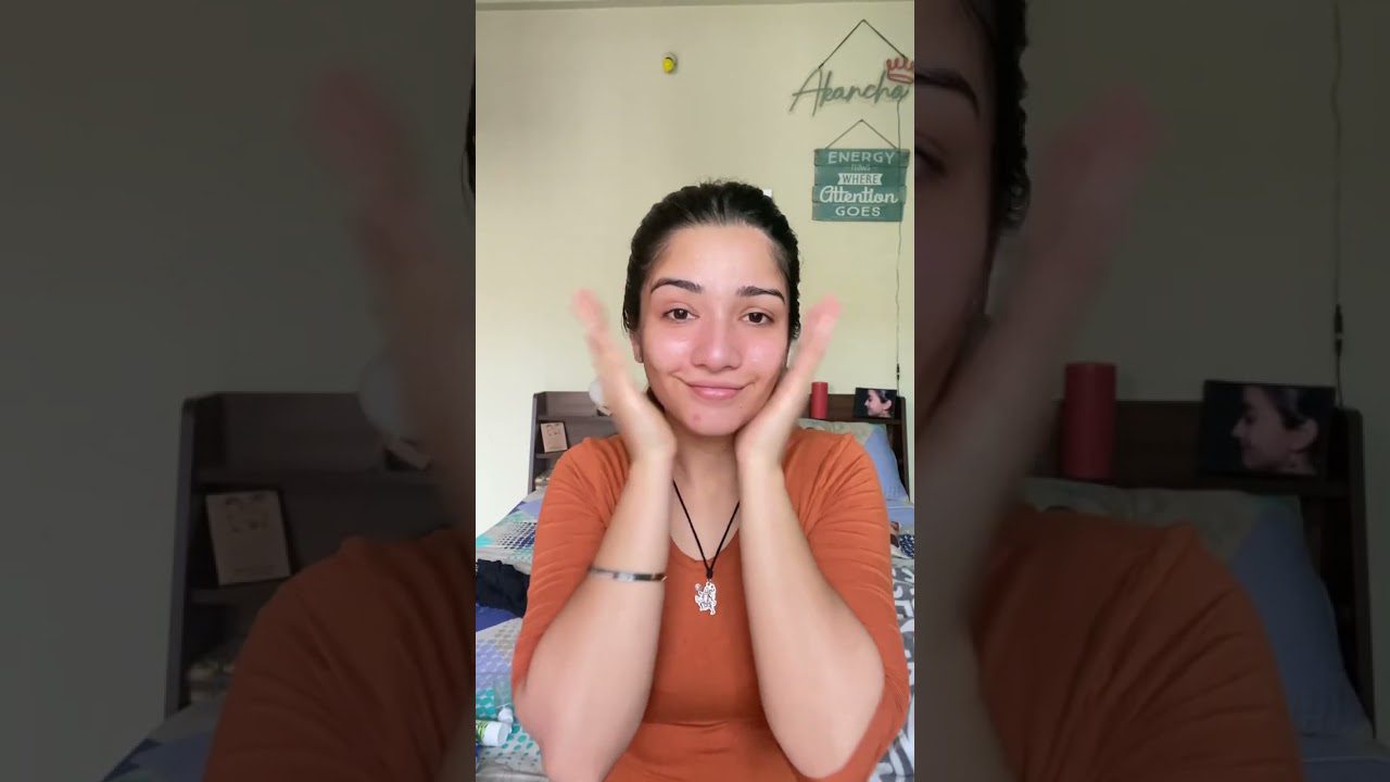 Jauna moteris oranžinėje palaidinėje šypsosi ir laiko rankas prie skruostų, kambaryje.