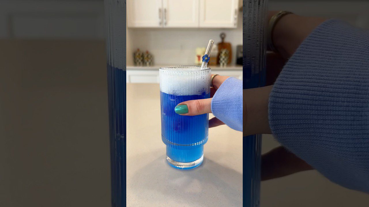 Rankus žmogaus delno laikomas aukštas stiklinis taurė su putojančiu mėlynu gėrimu.