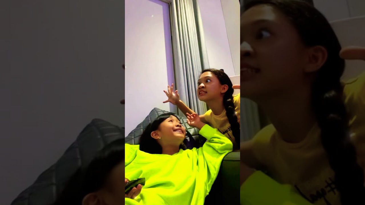 Dvi mergaitės ant sofos žaidžia ir juokiasi, viena laiko nuotolinio valdymo pultą.