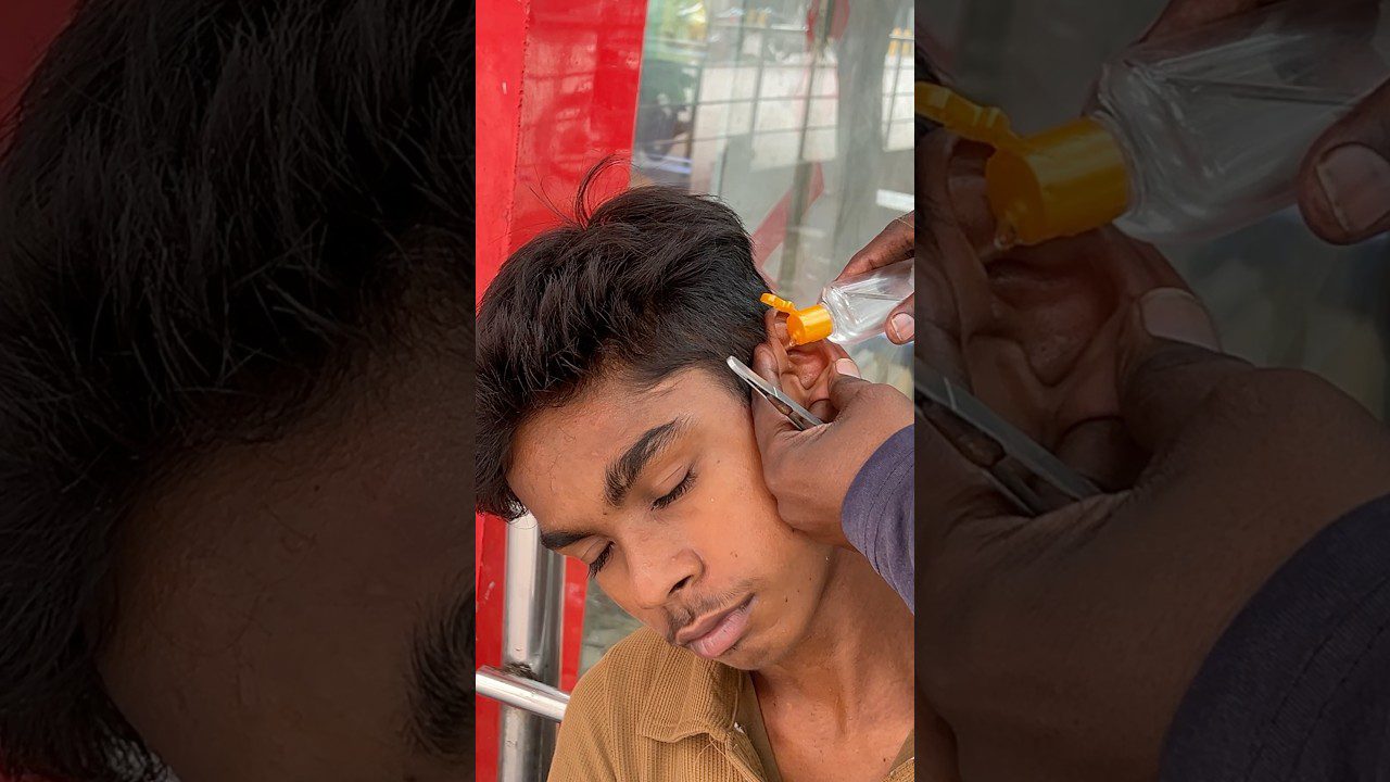 Jaunas vaikinas užmerktomis akimis, jam valoma ausis spaudžiant buteliuku ir žirklėmis.