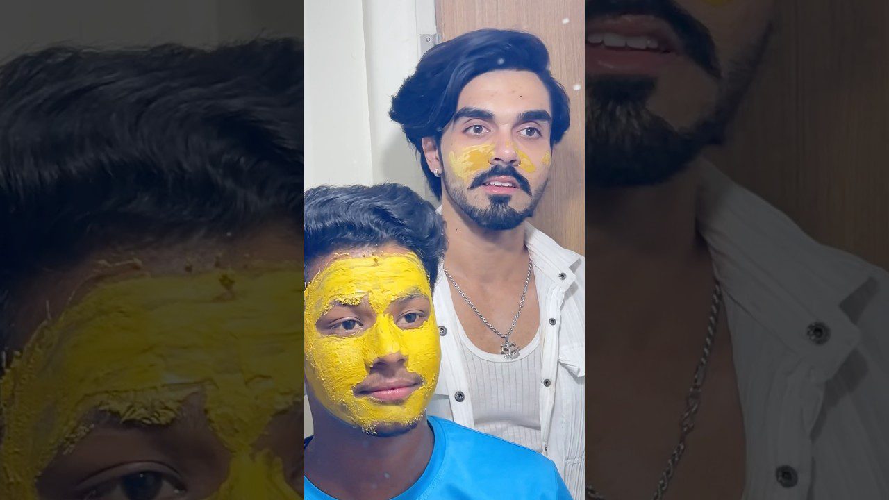 Du jauni vyrai veidais nutapytais geltonais kaukės pavidalo dažais, vienas sėdi, kitas stovi už jo.