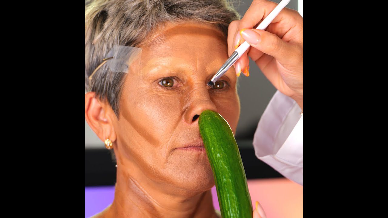 Moteris su trumpais pilkšvais plaukais, ant veido tepami makiažo potėpiai ir agurkas prie nosies