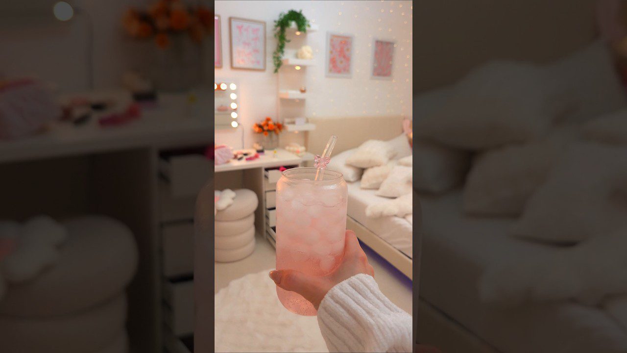 Ranka laiko stiklą rožinio ledo gėrimo priešais minkštai dekoruotą mergaitišką kambarį