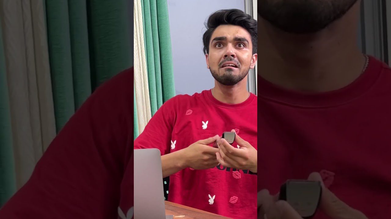 Jaunuolis raudonu marškinėliu nustebęs laiko išmanųjį laikrodį prie kompiuterio