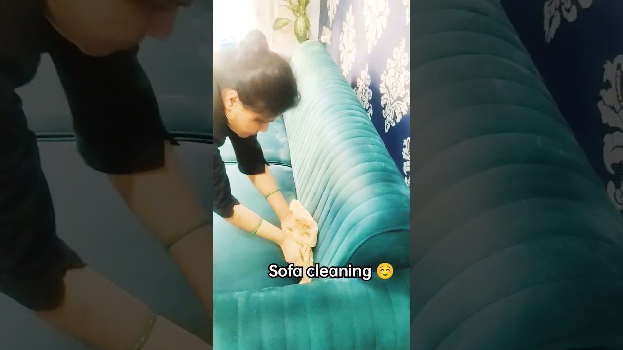 Moteris valo žalsvą velveto sofą rankšluosčiu, matomas užrašas „Sofa cleaning“ (sofos valymas).