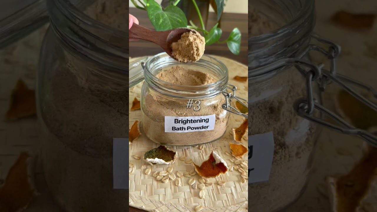 Stiklainis su „Brightening Bath Powder“ vonios milteliais, šaukštelis ir dribsniai bei apelsino žievelės