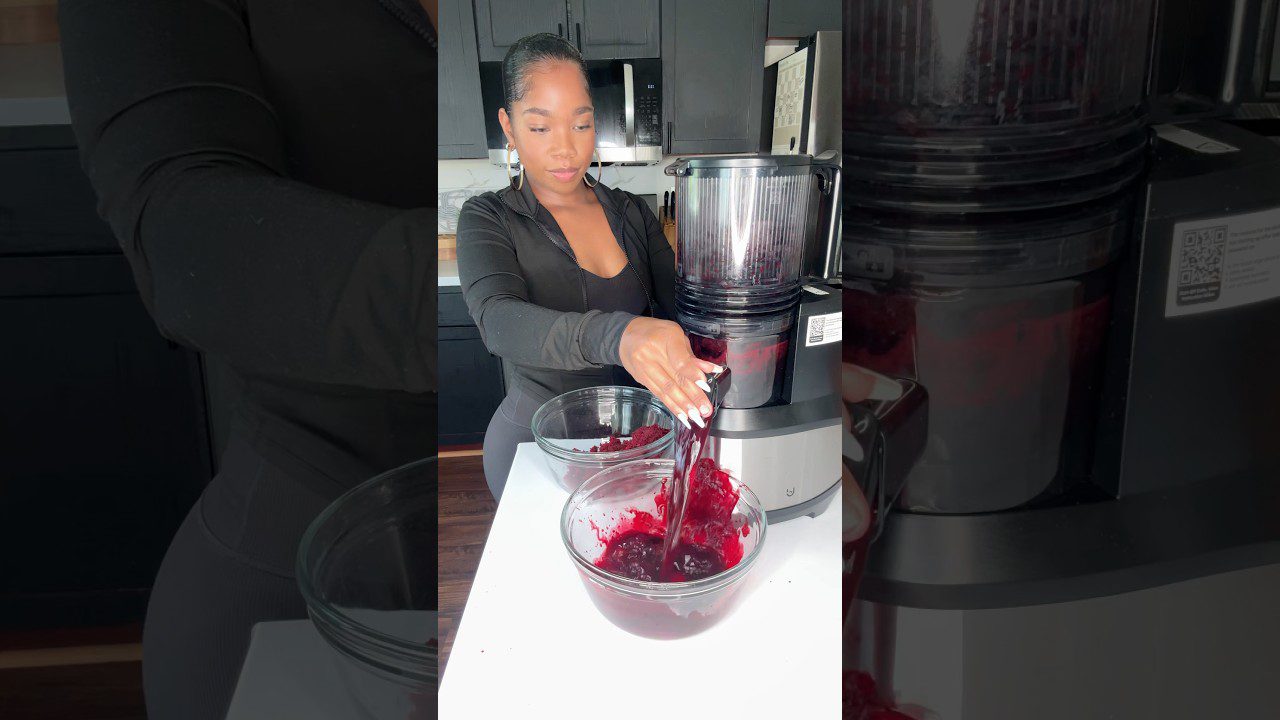 Moteris virtuvėje spaudžia raudonų uogų sultis į stiklinį dubenį su sulčiaspaude