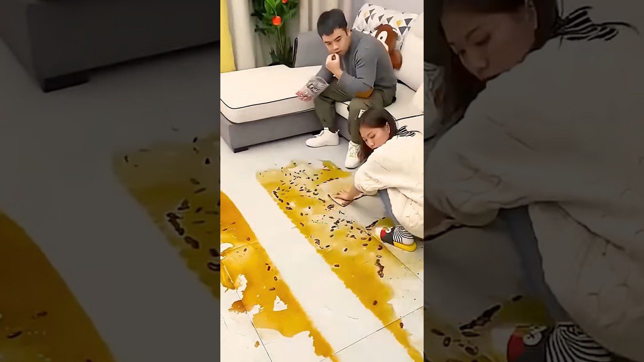 Moteris ant grindų valdo didelį geltoną išsiliejimą, vyro sėdint ant sofos stebi situaciją