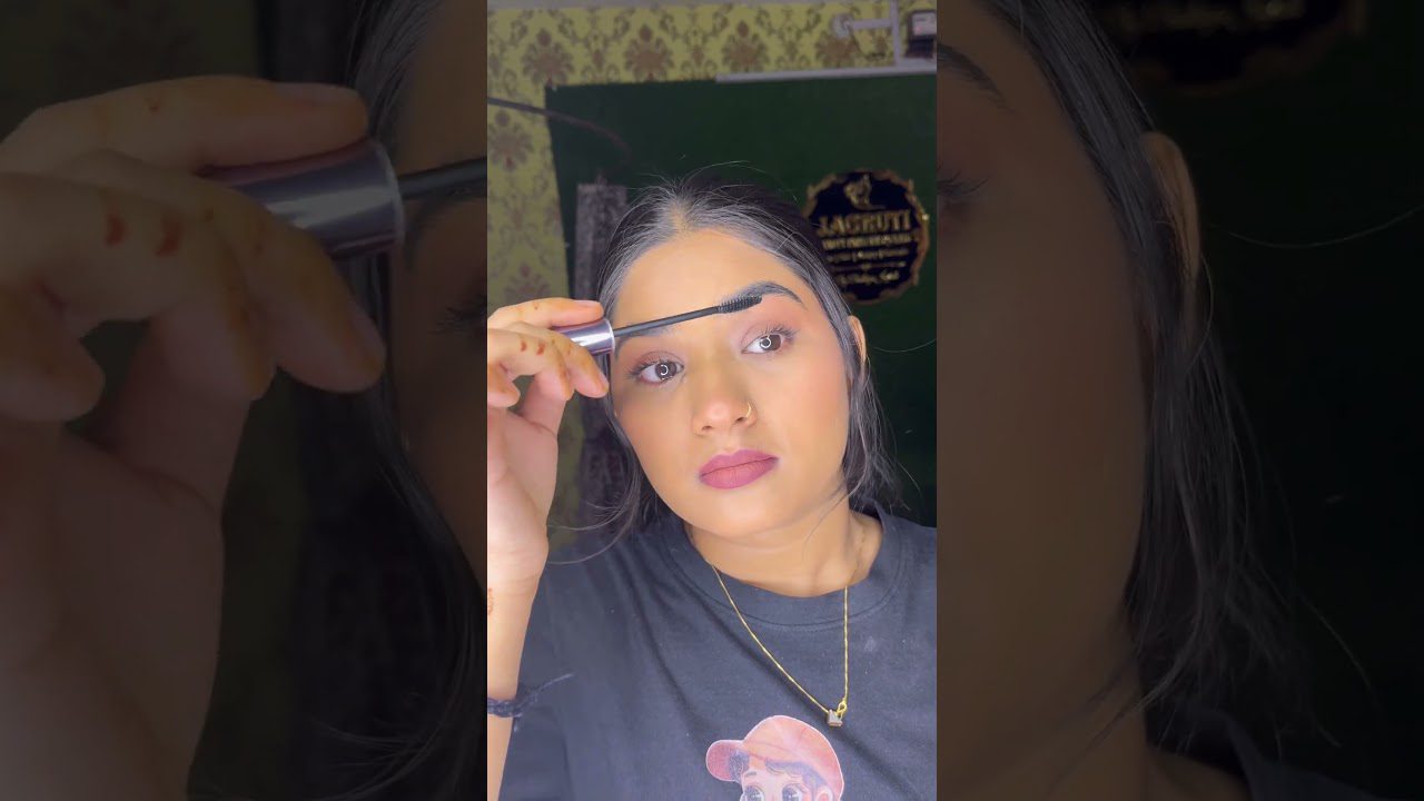 Moteris su tamsiais plaukais tepanti antakių gelį prieš veidrodį, dėvi aukso vėrinį.