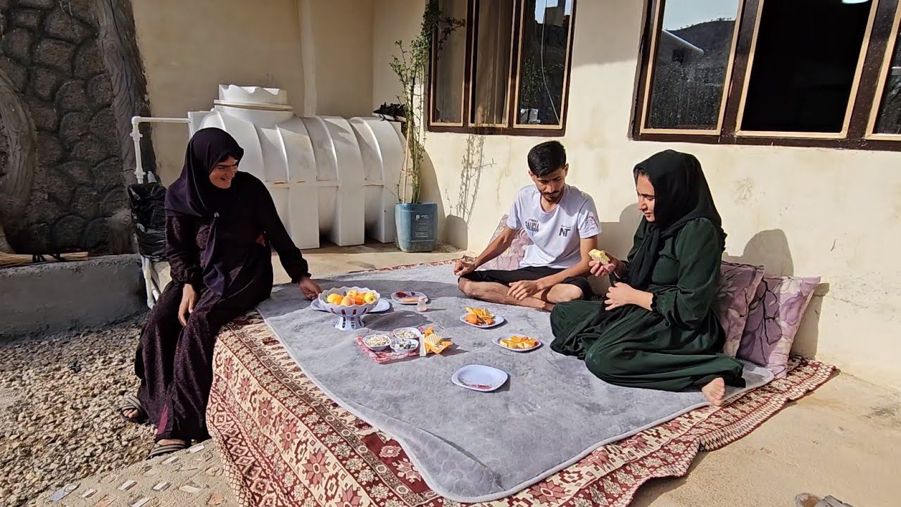 Trijų žmonių šeima sėdi ant kilimo lauko pavėsyje ir vaišinasi vaisiais bei užkandžiais.