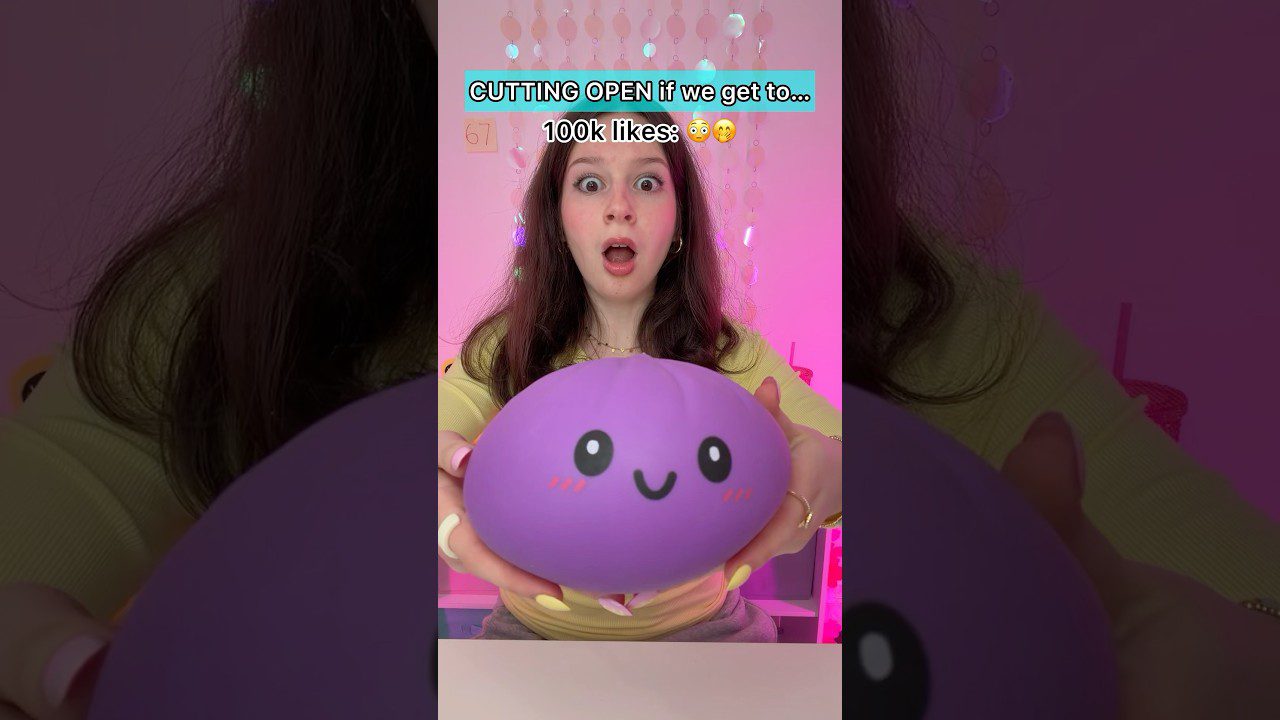 Jauna moteris nustebusi laiko violetinį šypseną turintį squishy žaislą prie kameros, viršuje užrašas „pjauti atidaryti“ (angl. cutting open).