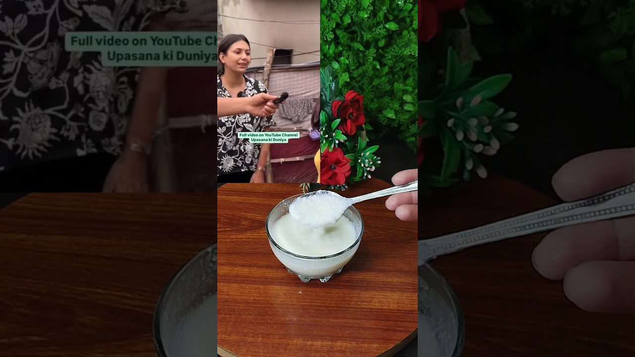 Stiklinė jogurto ant medinio stalo, šaukštas su kreminiu jogurtu virš jos, gėlės fone