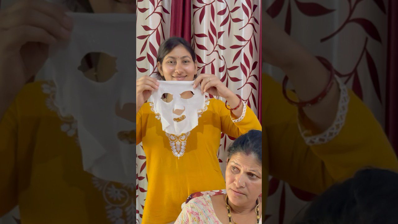 Moteris geltonais drabužiais laiko veido kaukę, šalia sėdi vyresnė moteris.