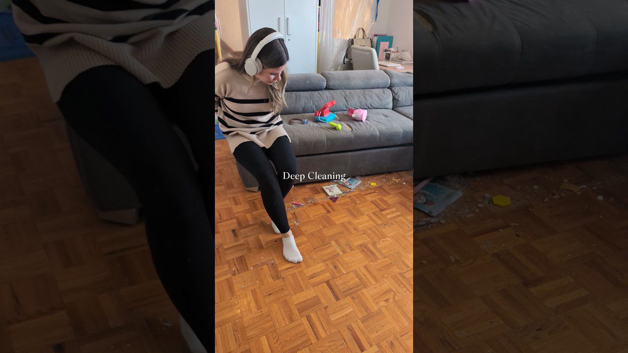 Moteris su ausinėmis sėdi ant krėslo šalia sofos, grindyse išmėtyti vaikų žaislai ir šiukšlės