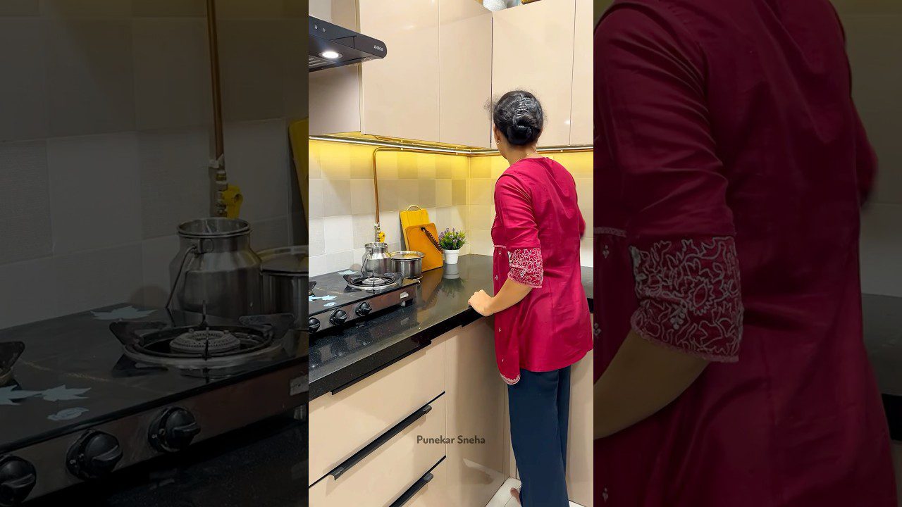 Moteris raudona palaidine stovinti prie virtuvės kaitlentės ir žiūrinti į viršutinius spinteles