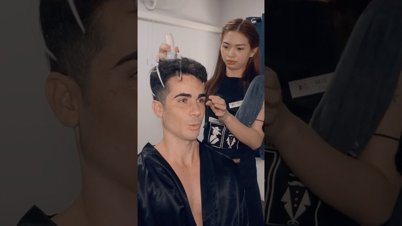 Vyras sėdi kirpykloje, stilistė purškia plaukus ir formuoja šukuoseną.