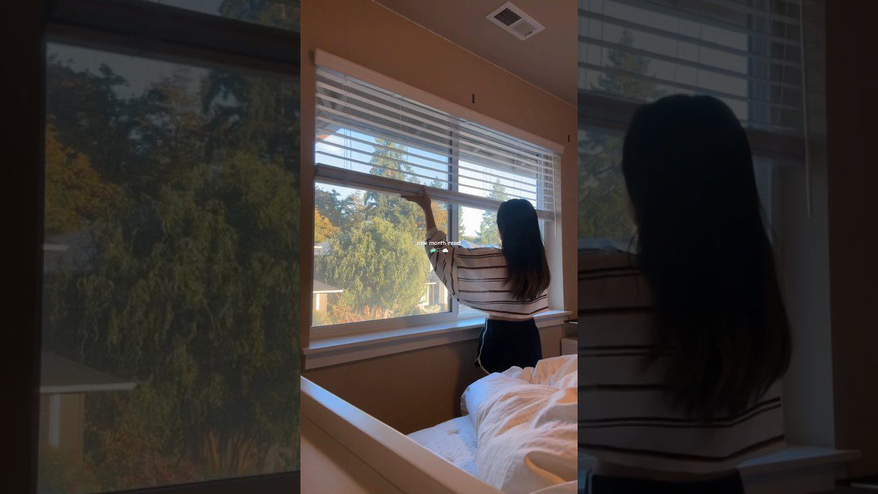 Moteris atidaro žaliuzes miegamojo lange, pro langą matyti medžiai ir namai