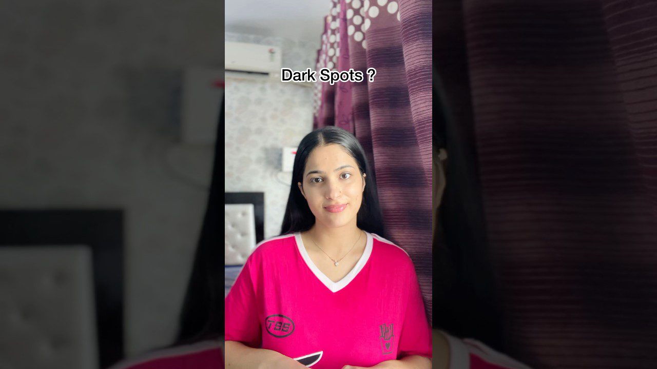 Jauna moteris su ilgais tamsiais plaukais dėvi rožinį marškinėlį ir šypsosi prie užuolaidų