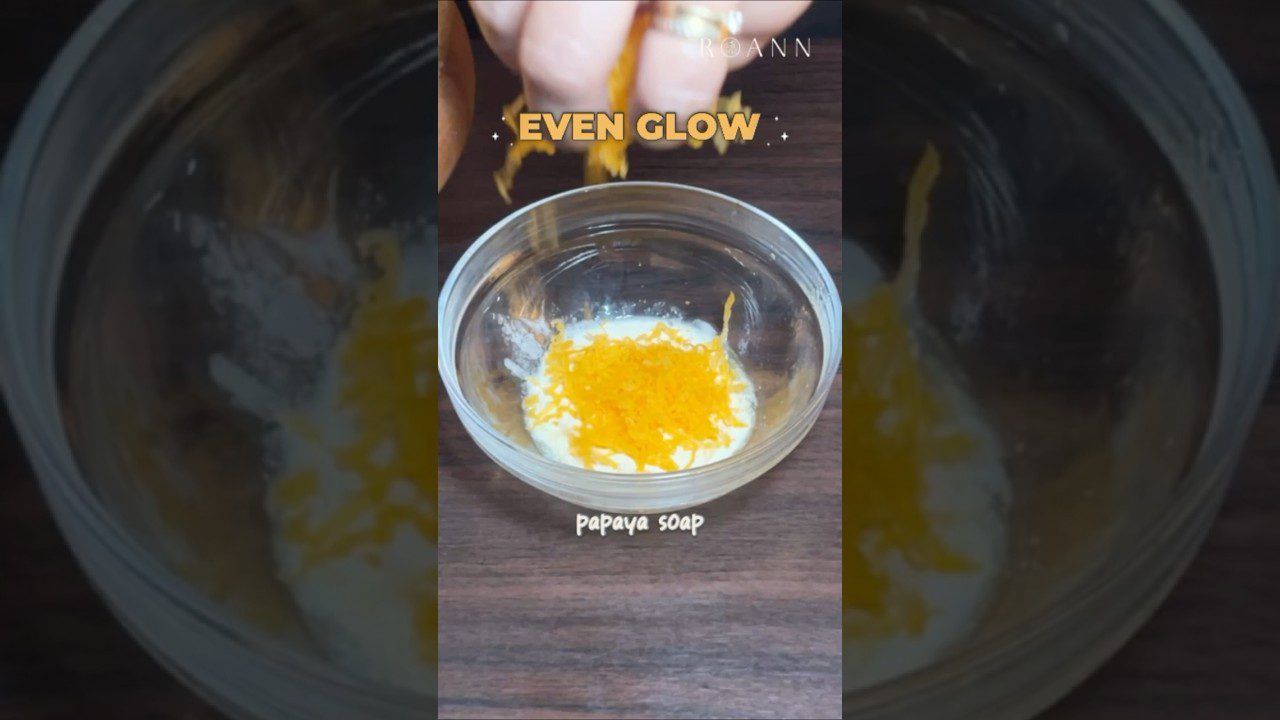 Stiklinėje dubenyje beriami papajos muilo drožlės ant kremo pagrindo, ranka viršuje.