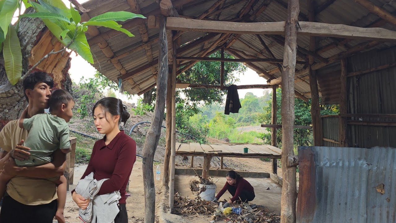 Jauna pora su vaikeliu prie atviros medinės trobėlės, moteris rūšiuoja malkas fone