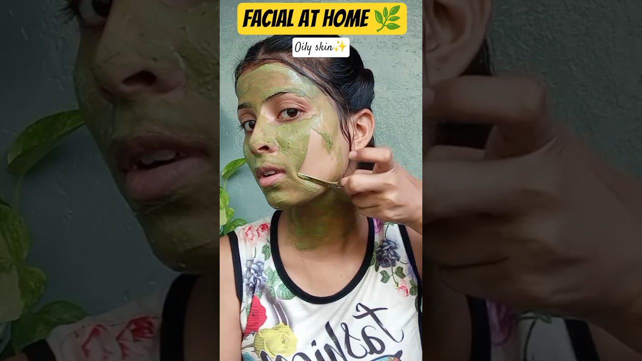 Moteris nuima žalią veido kaukę metaliniu įrankiu, užrašas „FACIAL AT HOME“ ir „Oily skin“