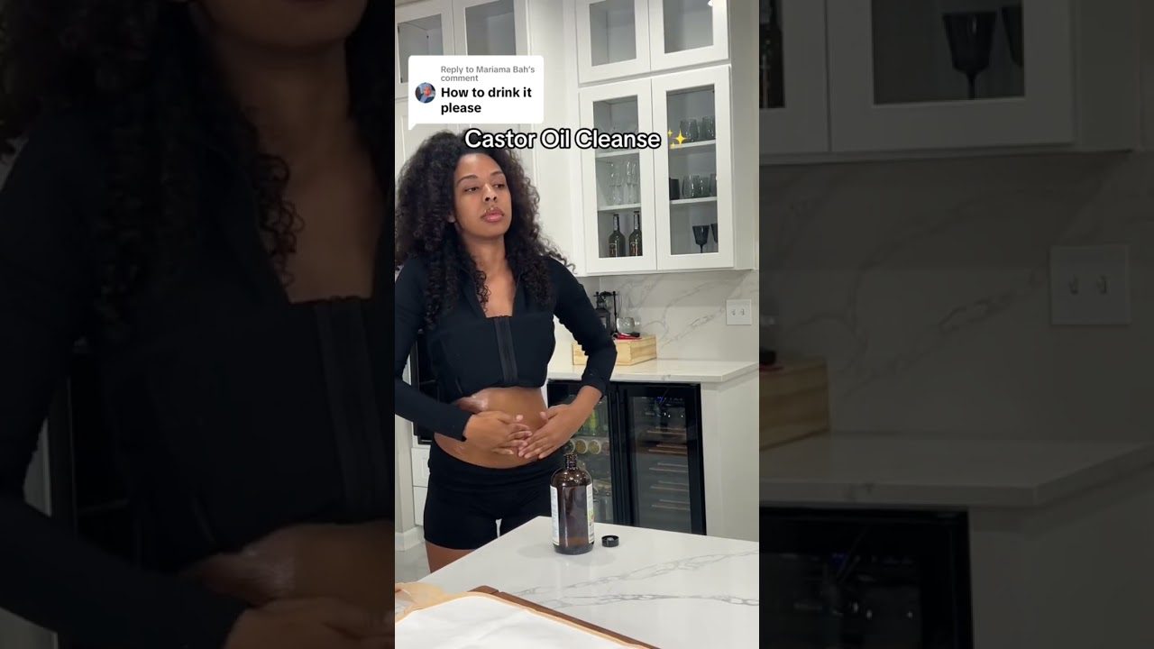 Moteris virtuvėje trinanti pilvą prie butelio su užrašu „Castor Oil Cleanse“