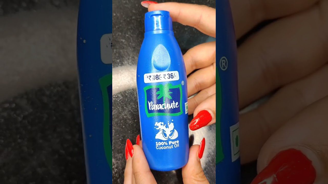 rankų laikoma mėlyna plastikinė Parachute kokosų aliejaus buteliukė su ryškiai raudonais nagais.