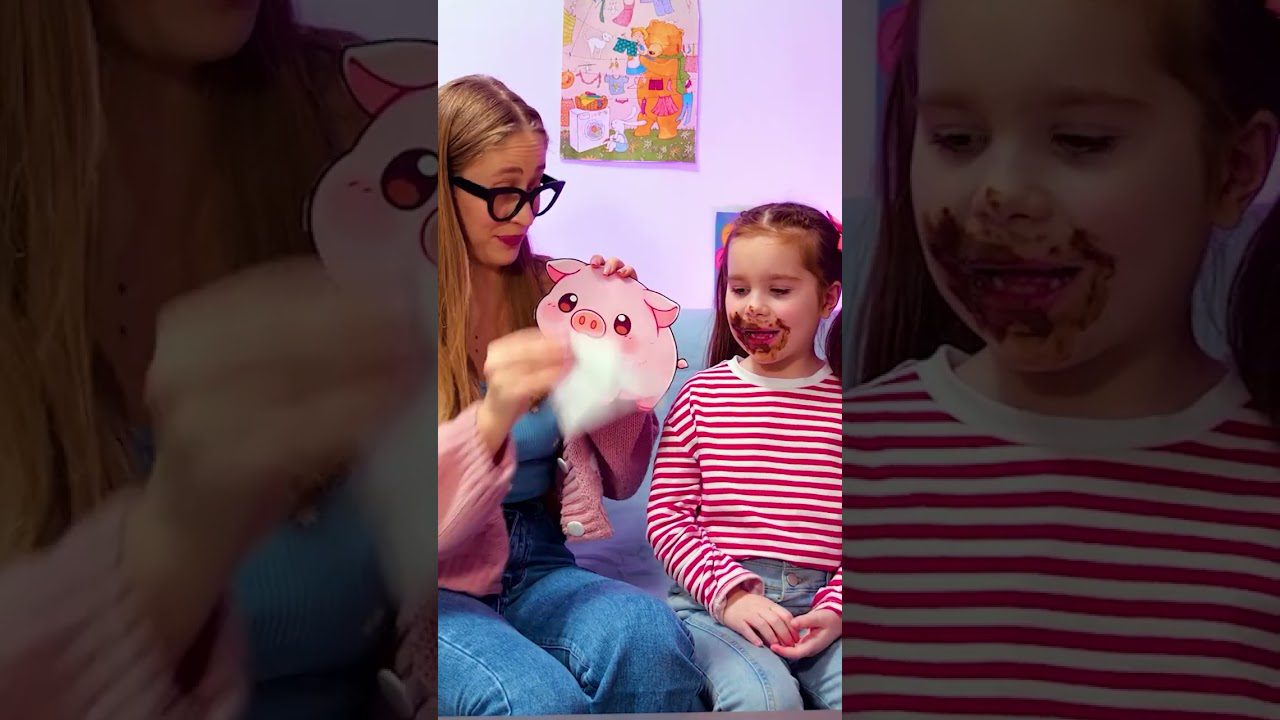 Moteris su akiniais valiko servetę, šalia sėdi mergaitė su šokoladu aplink burną.