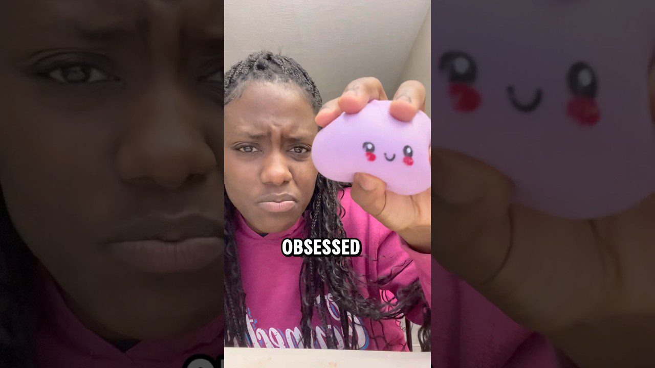 Jauna moteris laiko violetinį šypsenėlės formos minkštą žaislą, užrašas „OBSESSED“.