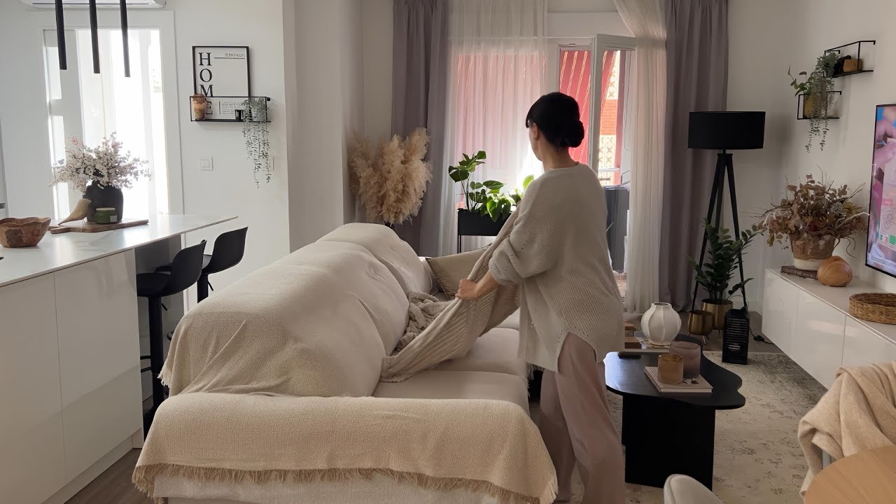 Moteris džiausto smėlio spalvos sofą šviesiame, augalų ir dekoracijų apstatytame svetainės kambaryje