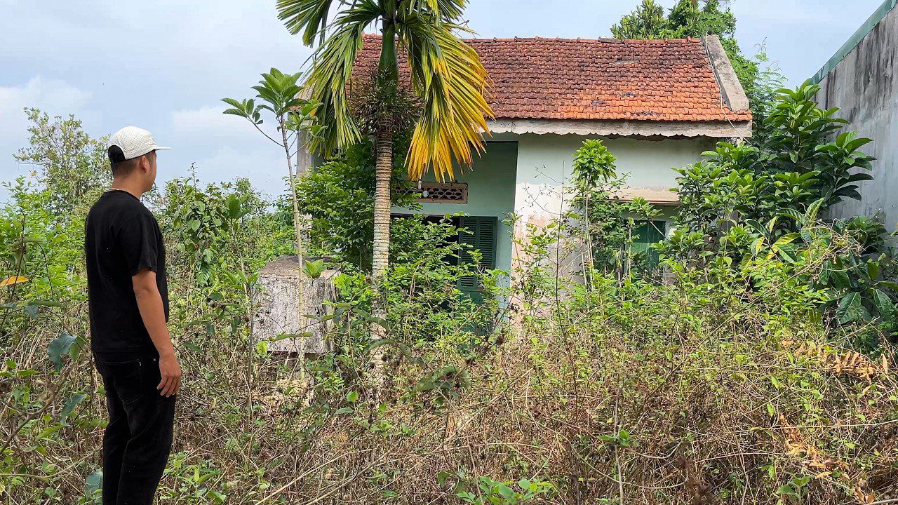 Vyras stovi prie apleisto namo, apsupto aukštų žolių ir medžių