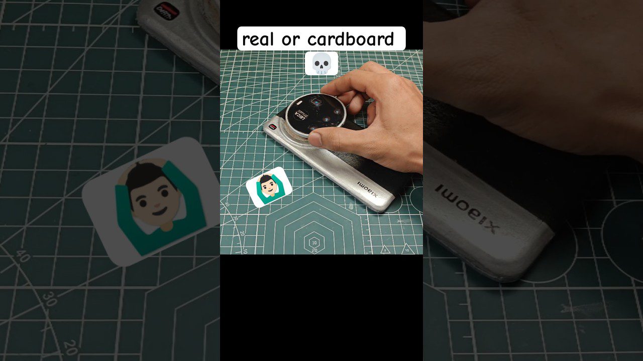 Rankos laiko kameros objektyvą virš pjovimo kilimėlio, šalia dviguba lipni plokštelė su veido piktograma ir užrašas „real or cardboard“.