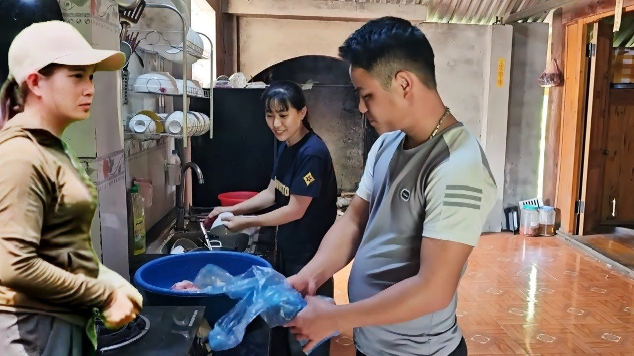 Trys žmonės virtuvėje plauna ir ruošia indus prie kriauklės ir stovinčio bako