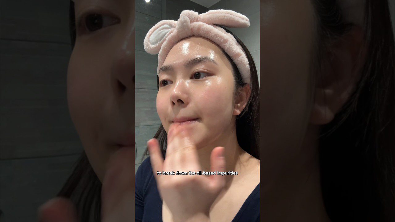 Moteris su rožiniu galvos juostos lanku tepa veido valymo priemonę, matomas subtitras apie riebalines priemaišas