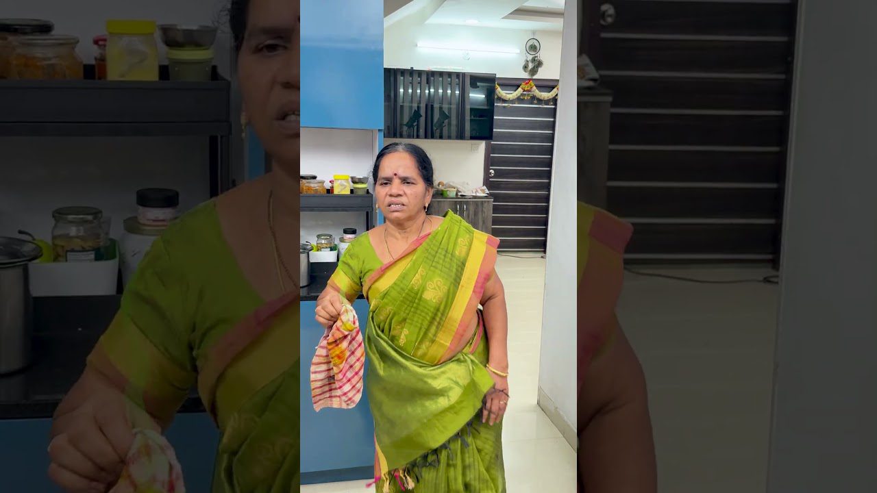 Moteris vilkinti žalia sari virtuvėje laiko skudurėlį ir žiūri į kamerą.