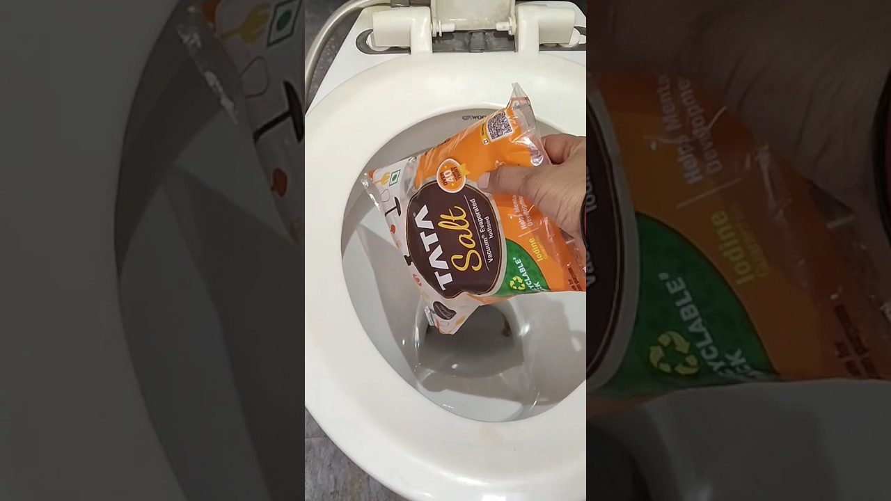 Rankoje laikomas pakelis TATA druskos virš atidaryto tualeto dubens.