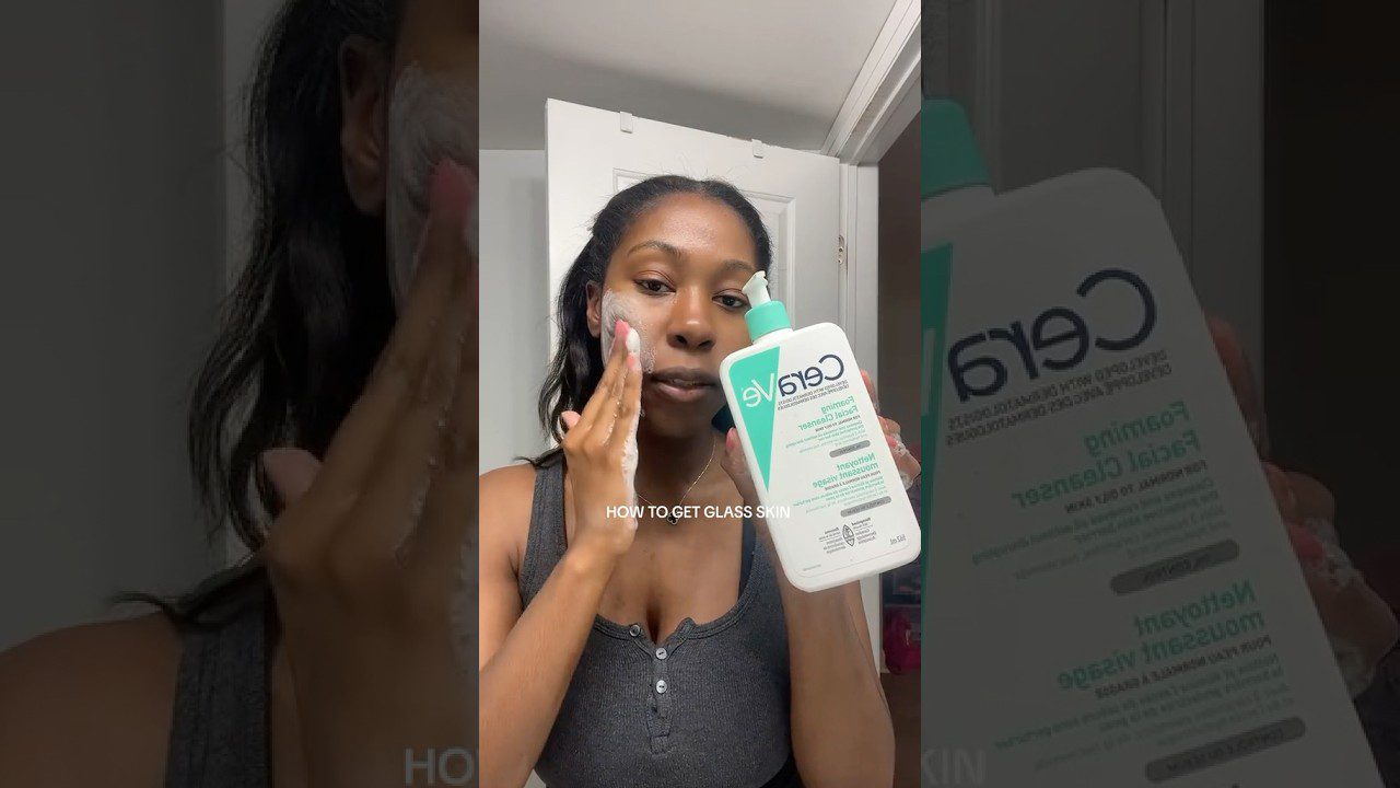 Moteris veidą plauna putomis ir laiko CeraVe valomąjį gelį su užrašu „Foaming Facial Cleanser“.