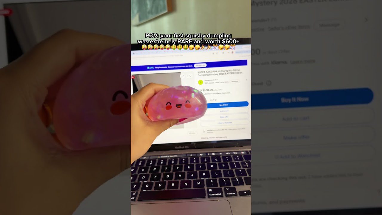 Rankoje laikomas rožinis blizgantis squishy kramtukas su šypsena priešais atidarytą kompiuterį.