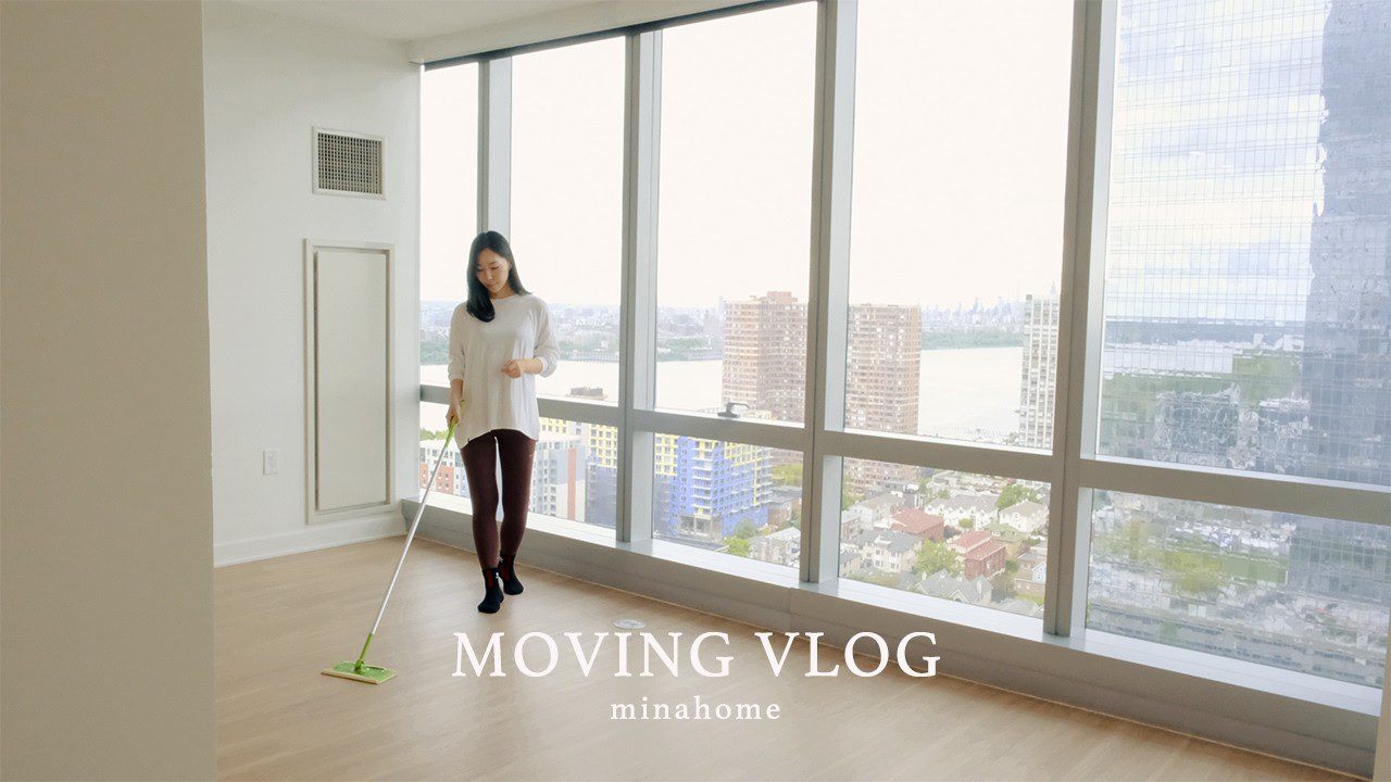 Moteris valo tuščius apartamentus prie didelių langų su miesto vaizdu ir užrašu "Moving Vlog"