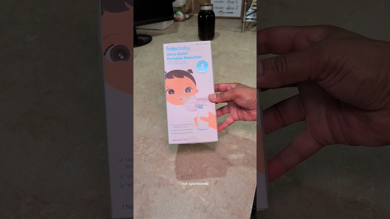 rankose laikomas Frida Baby tylus nešiojamas nebulizatorius su pakuotės iliustracija ir užrašu "Ultra-Quiet Portable Nebulizer"