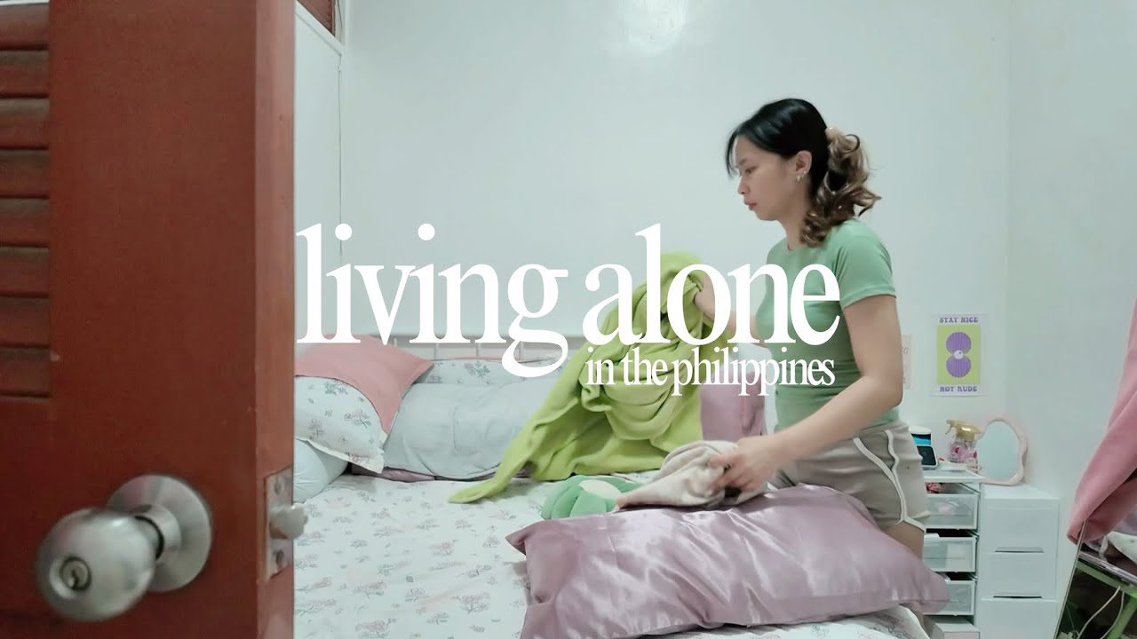 jauna moteris tvarko lovą mažame kambaryje, ant sienos užrašas „living alone“ ir „in the philippines“