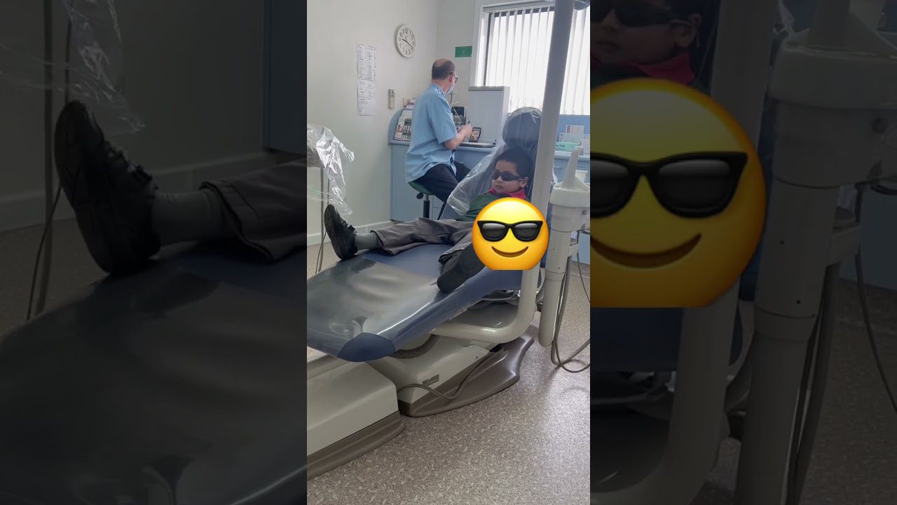 Vaikas su akiniais gulintis odontologinėje kėdėje, fone dantų gydytojas prie įrangos