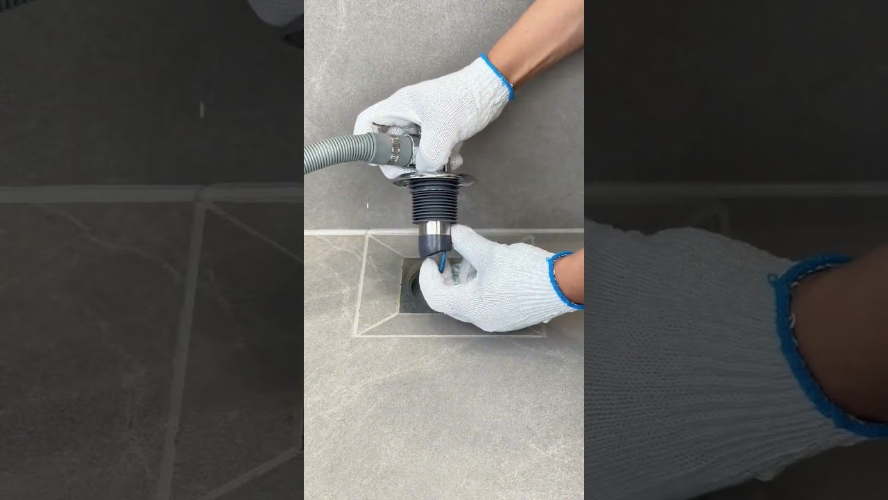 Rankos su darbo pirštinėmis montuoja lankstų latako vamzdį į grindų nutekėjimą.