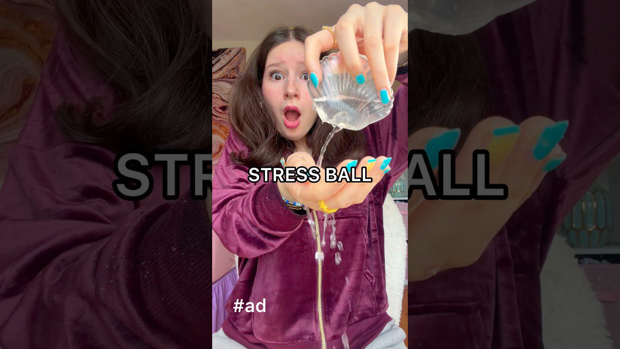 Jauna moteris lieja gelsvą skysčio rutulį rankoje, užrašas „STRESS BALL“.