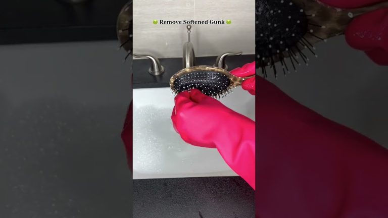 Rankos su rožiniais guminėmis pirštinėmis valo plaukų šepetį virš kriauklės, matomas užrašas "Remove Softened Gunk".