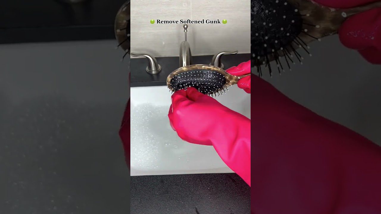 Rankos su rožiniais guminėmis pirštinėmis valo plaukų šepetį virš kriauklės, matomas užrašas "Remove Softened Gunk".