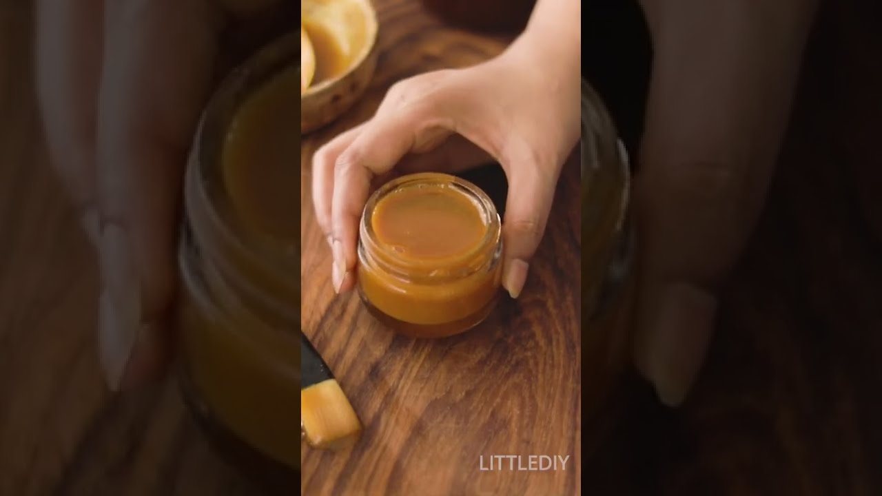 Rankos laiko mažą stiklainį su kreminiu karamelės arba medaus spalvos tepalų mišiniu ant medinio paviršiaus.